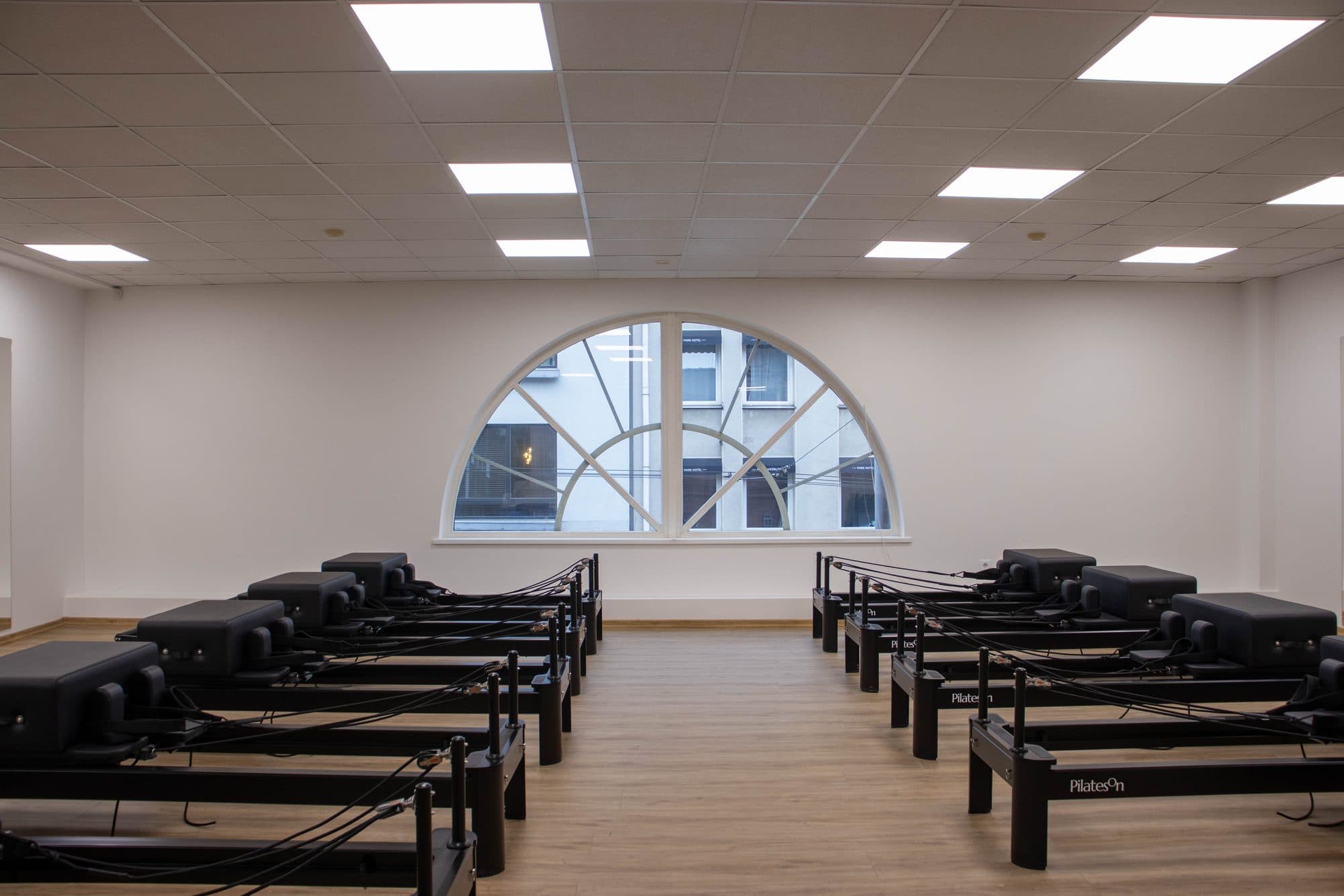 Šviesi, simetriška „PilatesOn“ reformerių salė su reformerių eilėmis prieš pusmėnulio formos langą